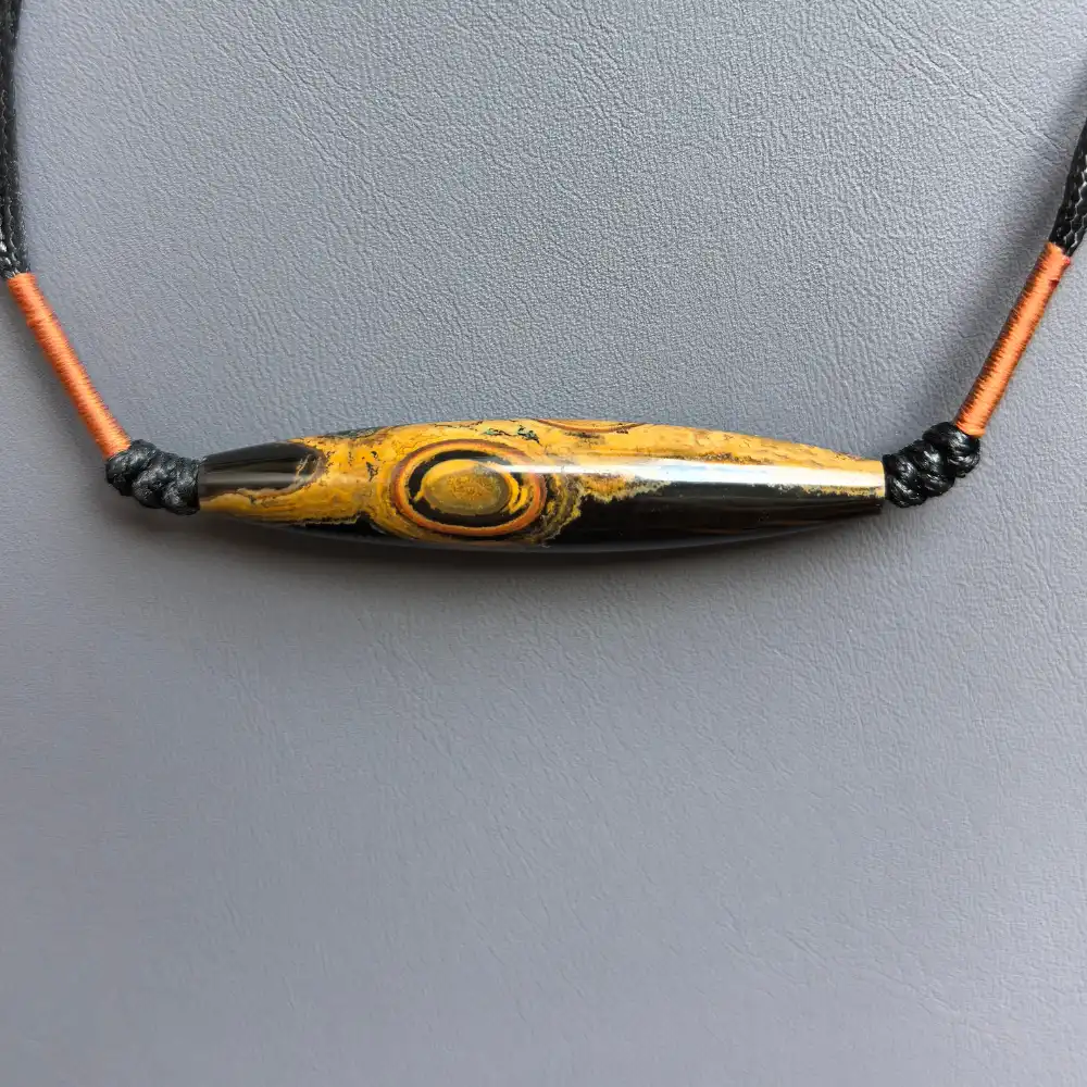 Flat-lay of the two-eye eye-stone lance pendant on grey texture with black cord | Glint & Grain