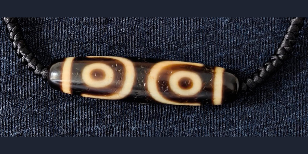 Three-eyed dzi bead strung on a hand-knotted black cord, shown against a dark textured background | Glint & Grain