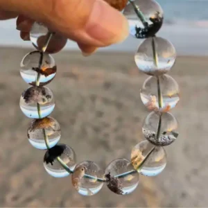 Hand cradling a bracelet of clear quartz beads with black and rust-brown phantom inclusions against a blurred beach background