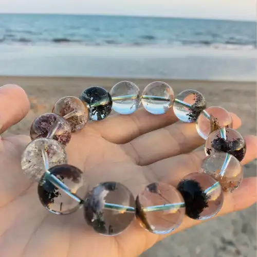 Hand cradling a bracelet of clear quartz beads with black and rust-brown phantom inclusions against a blurred beach background