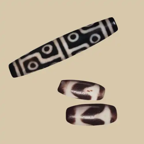 Two Dzi beads on a beige background — a long multi-eye bead with dark line motifs, above a tiger-tooth Dzi shown from the front and the back.